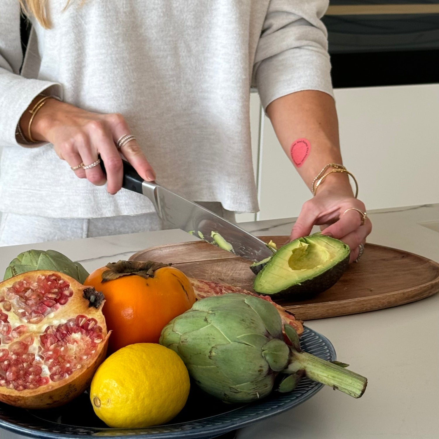 Person cutting an avocado on a wooden cutting board with various fruits and vegetables on a table, showing a GLP-1 patch sticked to the arm.